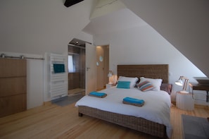 Quarto com cama grande, mesa de cabeceira com abajur, guarda-roupa de madeira e banheiro visível por uma porta aberta.