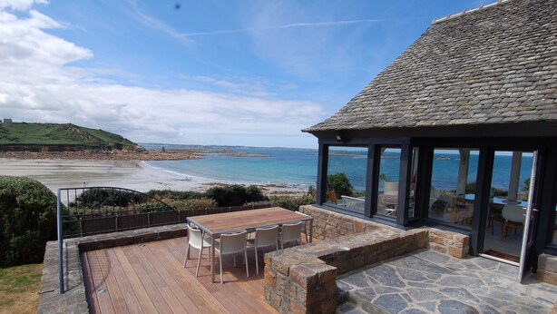 Eine Holzterrasse mit Tisch und Stühlen, ein Steinkamin und eine Glaskonstruktion mit Blick aufs Meer.
