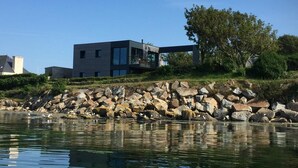A modern house by a lake with a rocky shoreline and a tree.