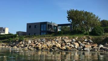 Une maison moderne située au bord d’un lac, avec une rive rocheuse et un arbre.