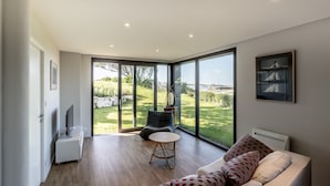 A spacious living room with a large sliding glass door leading to a garden, a comfortable sofa, a small round table, and a flat-screen TV on a white stand.