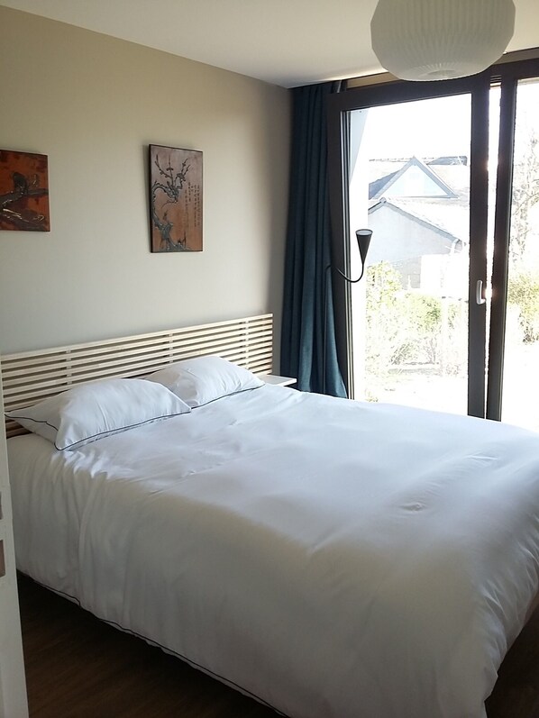 Una cama bien tendida con sábanas blancas en una habitación con cabecera blanca, una ventana con cortinas y dos cuadros colgados en la pared.