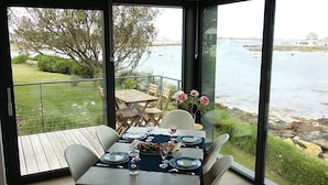 A dining area with a table set for four, overlooking a body of water with boats and a rocky shoreline.