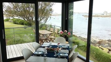 Un coin repas avec une table dressée pour quatre personnes, donnant sur une étendue d’eau où l’on peut voir des bateaux et une côte rocheuse.