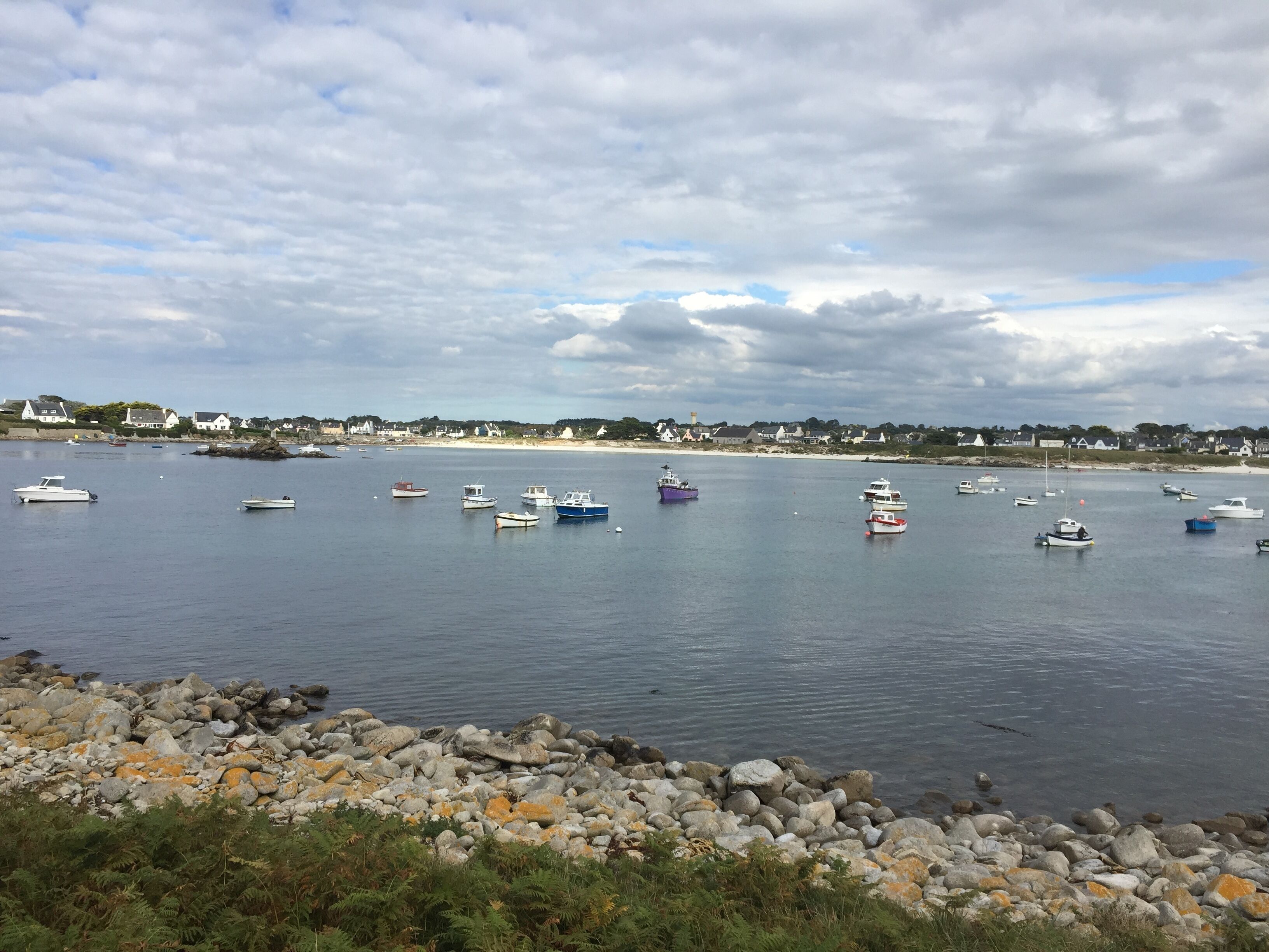 Escena costera con barcos en aguas tranquilas, un litoral rocoso y casas al fondo.