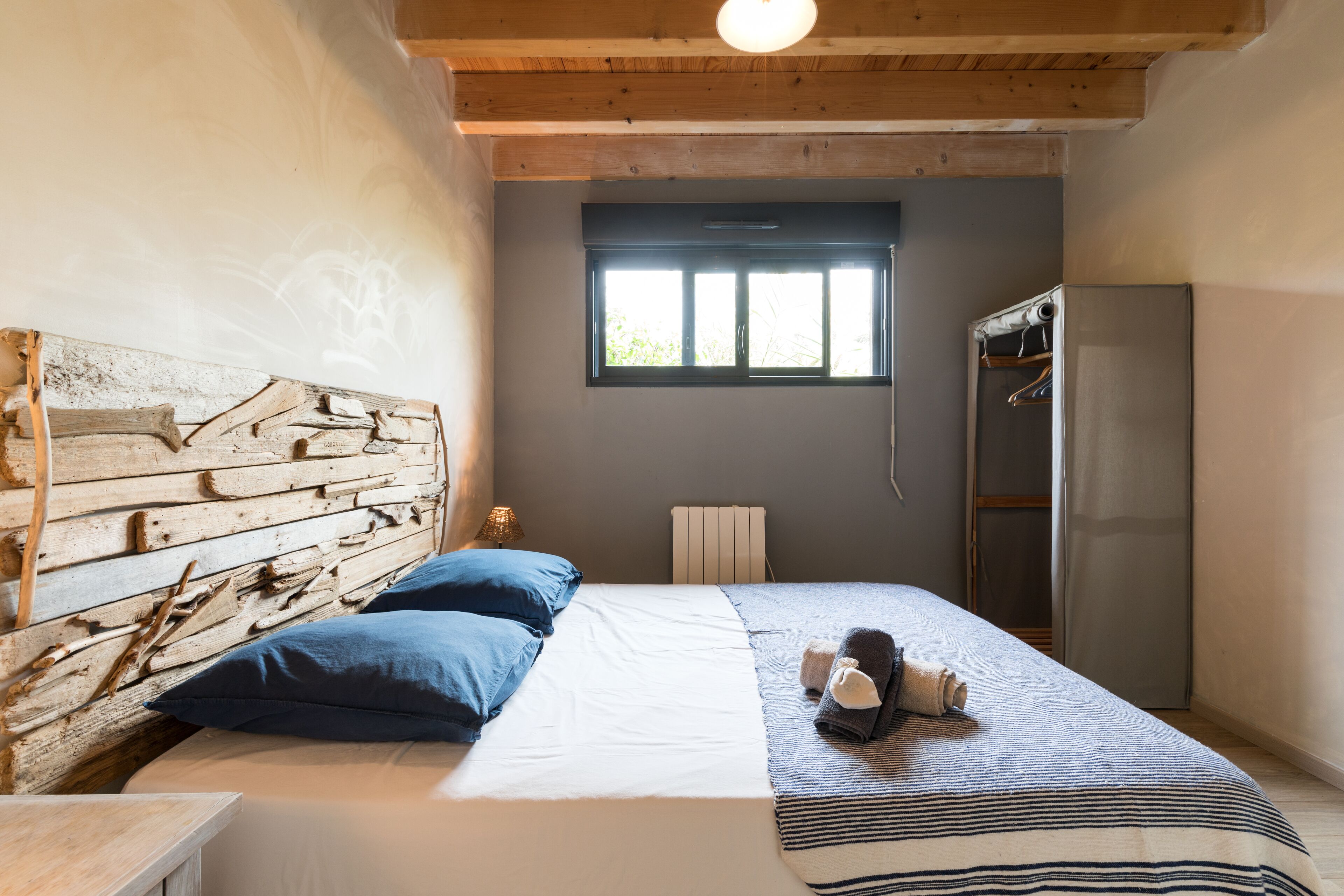 Ein Schlafzimmer mit einem hölzernen Kopfteil, einem Bett mit weißer und blauer Bettwäsche, einem Fenster und einem Kleiderschrank.