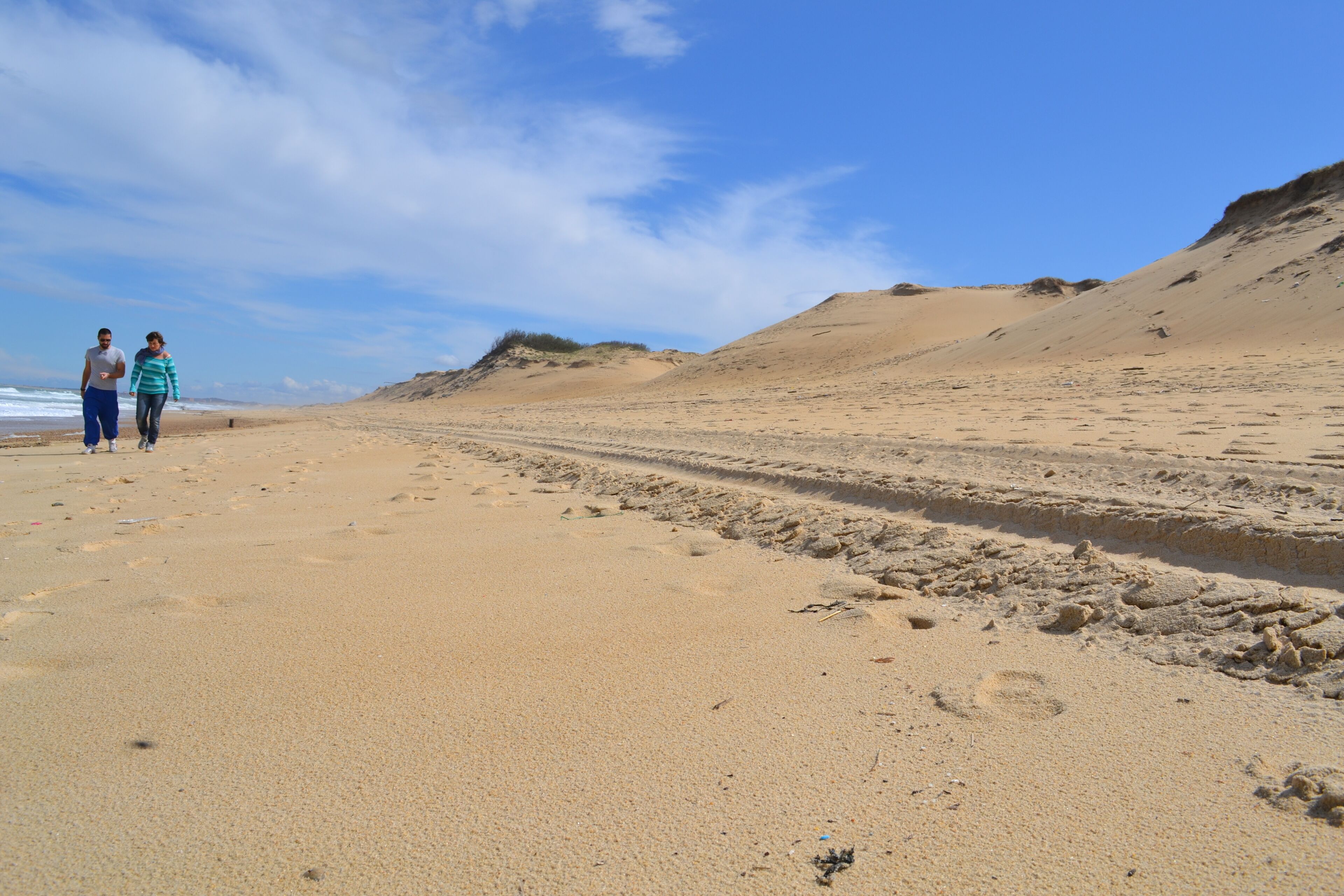 Ein Sandstrand mit Fußabdrücken und Radspuren, zwei Personen beim Spaziergang, und Dünen im Hintergrund.