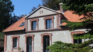 Une maison à deux étages, avec un toit recouvert de tuiles rouges, une façade en briques et un petit jardin agrémenté d’une plante en pot.