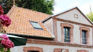 Une maison à deux étages, avec un toit recouvert de tuiles rouges, des murs en briques et un petit jardin agrémenté de chaises et d’une table, le tout de couleur blanche.