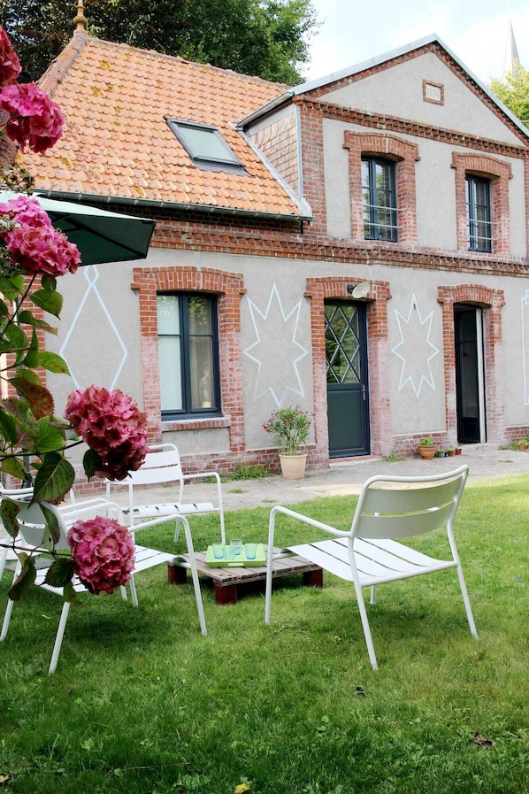 Une maison à deux étages, avec un toit recouvert de tuiles rouges, des murs en briques et un petit jardin agrémenté de chaises et d’une table, le tout de couleur blanche.