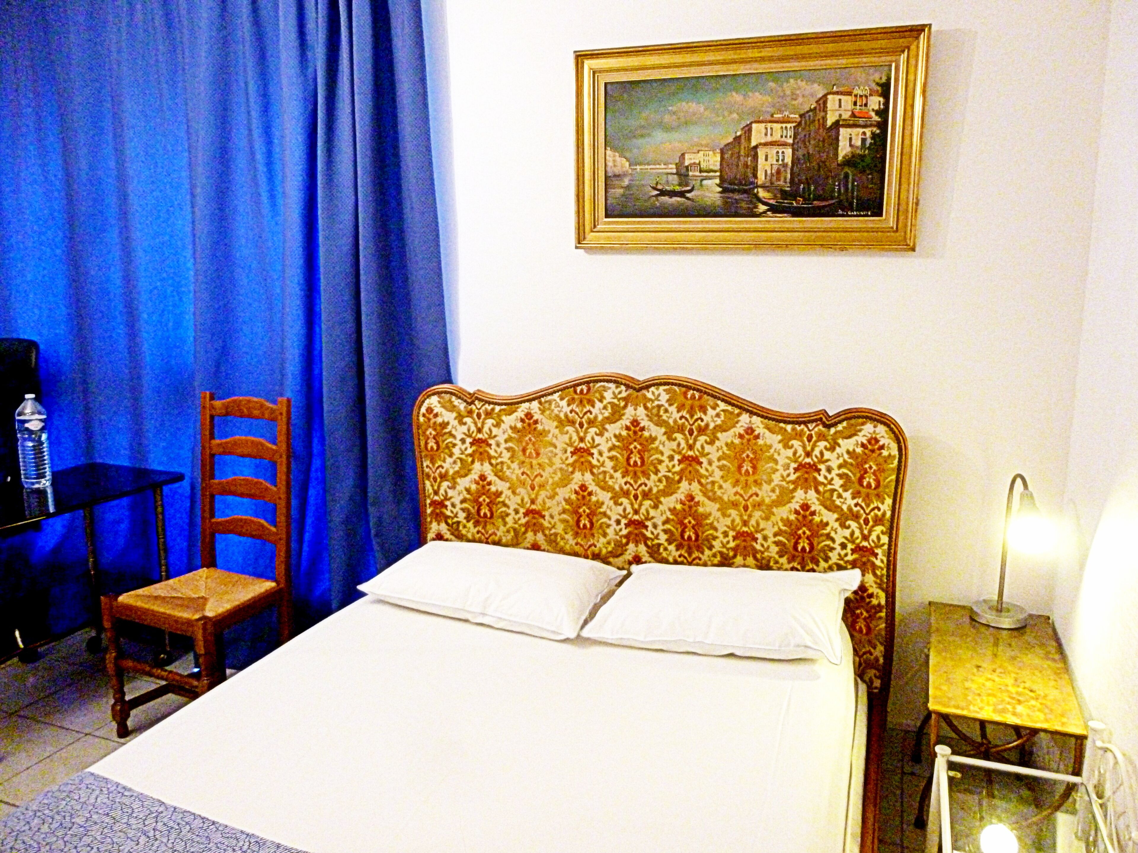 A hotel room with a bed, a chair, a table, a lamp, and a painting on the wall.