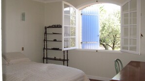 A bedroom with a bed, a chair, a wooden shelf, and a window with a blue door. - villa, countryside, quiet, spacious (8 persons), secure swimming pool. (Sérignan-Du-Comtat)