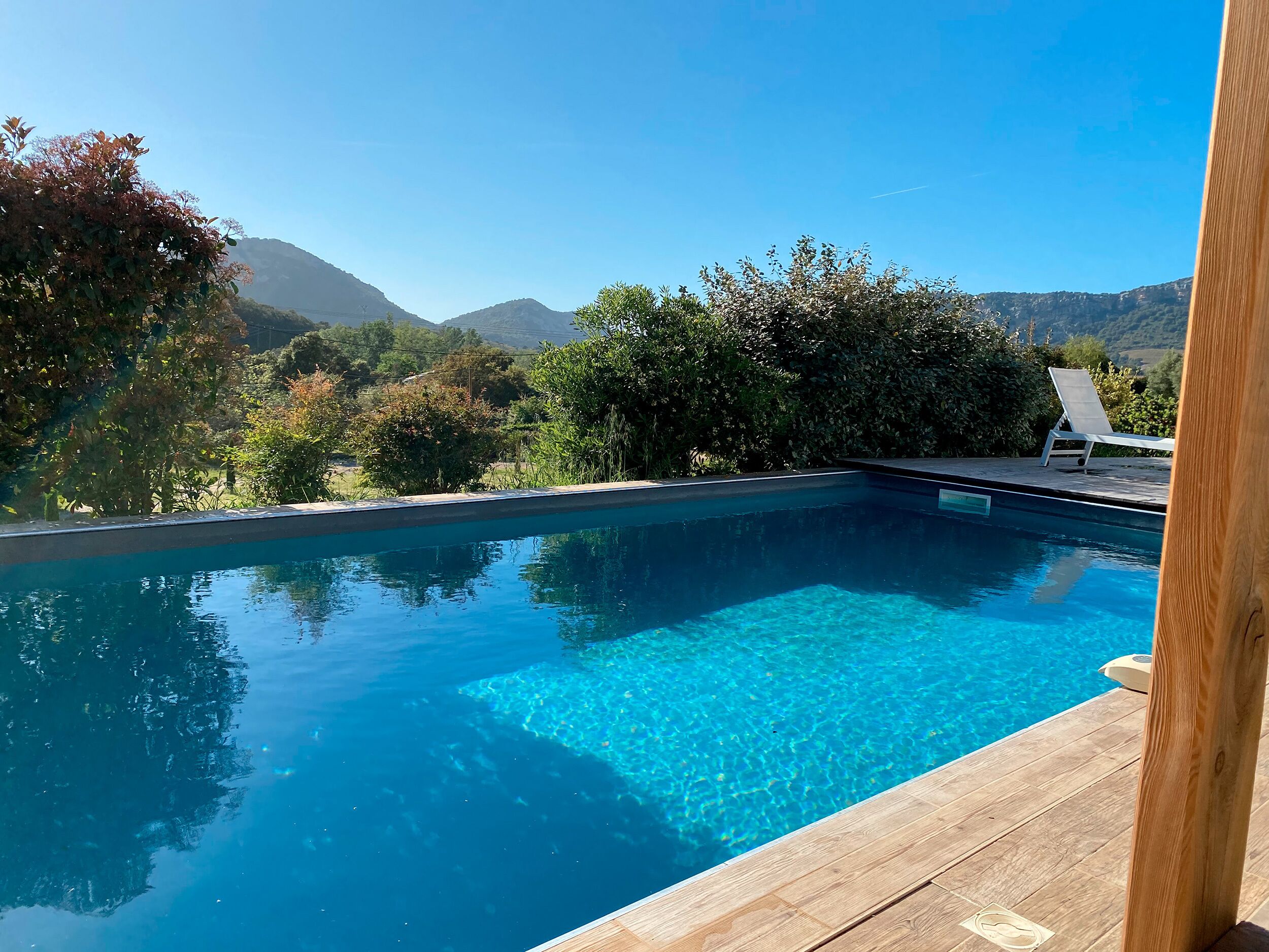 En klar, blå pool med en træterrasse, omgivet af frodig vegetation og bjerge i baggrunden.