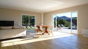 A spacious living room with a large sofa, a dining table with chairs, and a sliding glass door leading to an outdoor area with a pool and mountain view. - New villa with swimming pool overlooking a hectare of land overlooking the vineyard (Patrimonio)