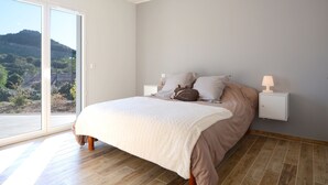 A bedroom with a large bed, a nightstand, a lamp, and a view of the outdoors. - New villa with swimming pool overlooking a hectare of land overlooking the vineyard (Patrimonio)