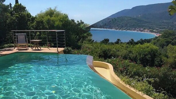 A clear pool with a slide, surrounded by greenery and overlooking a coastal landscape.