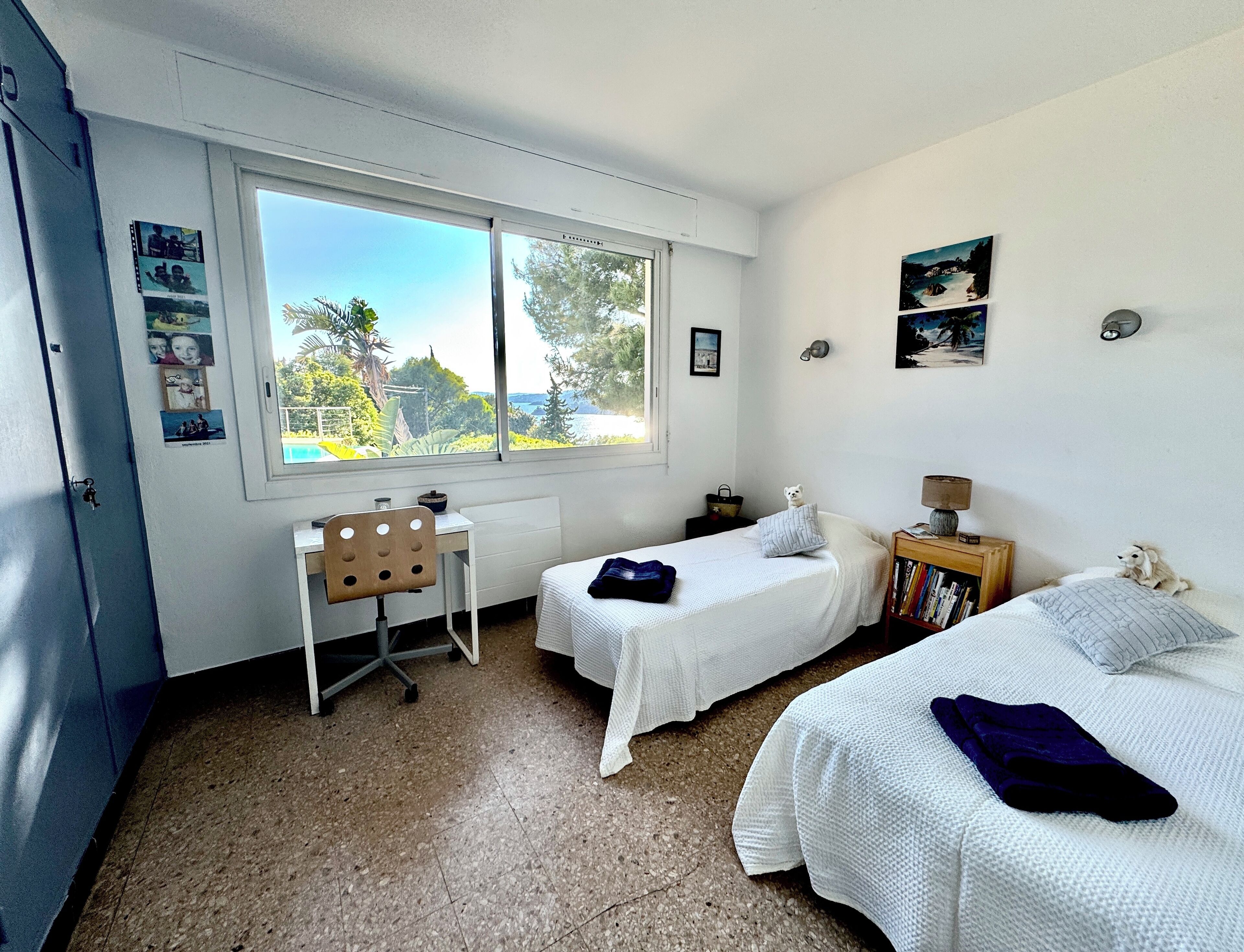 A bedroom with two beds, a desk, and a window with a view of trees and a body of water.