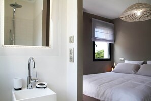 A modern bathroom with a shower, sink, and toilet. There is a bed with white bedding and a hanging light fixture.
