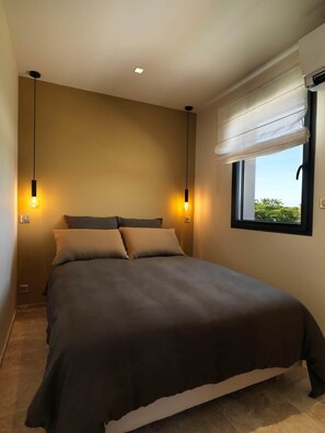 A bedroom with a bed, two wall-mounted lights, a window with a blind, and a view of greenery outside.