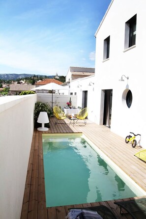 A rooftop pool with wooden decking, a dining area with yellow chairs, and a white building with a circular window.