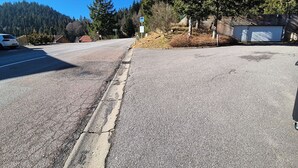Une route avec une voiture blanche garée sur le côté gauche, un trottoir endommagé et une colline boisée en arrière-plan.