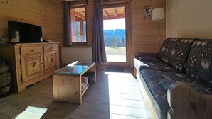 Un intérieur de chalet chaleureux avec un plancher en bois, un canapé orné de motifs en forme de cœur, un meuble en bois doté de poignées en forme de cœur, une table basse avec un plateau en verre et un téléviseur posé sur un meuble.