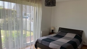 A bedroom with a bed, a nightstand, a lamp, and a view of the outdoors through sheer curtains. - Apartment T3 in Larmor beach 5 minutes walk from the beaches (Larmor-Plage)