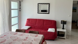 A room with a red sofa, a dining table with a patterned tablecloth, a mirror, and a lamp on a small table. - Apartment T3 in Larmor beach 5 minutes walk from the beaches (Larmor-Plage)