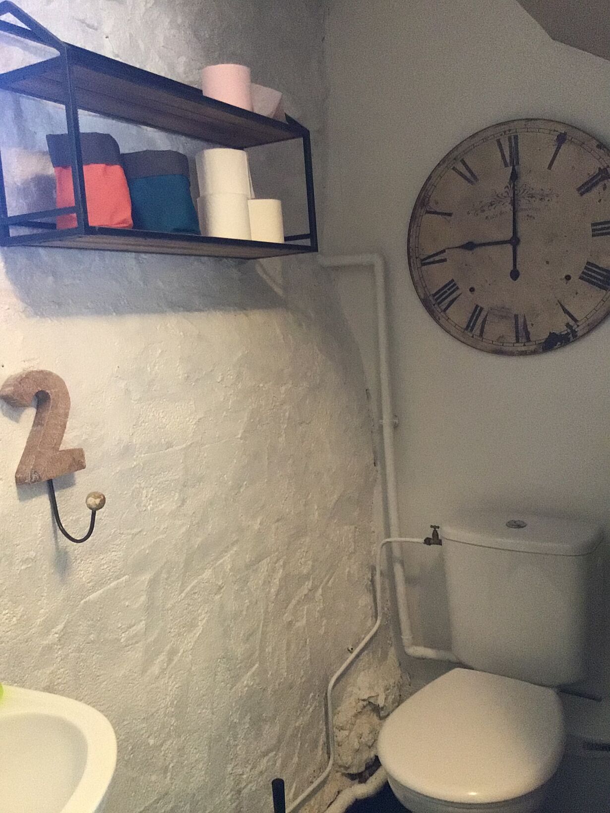 Ein Badezimmer mit Toilette, Waschbecken, einer Uhr und einem Regal mit Toilettenpapier und weiteren Gegenständen.