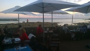 An outdoor dining area with tables and umbrellas overlooking a rocky beach and the sea.