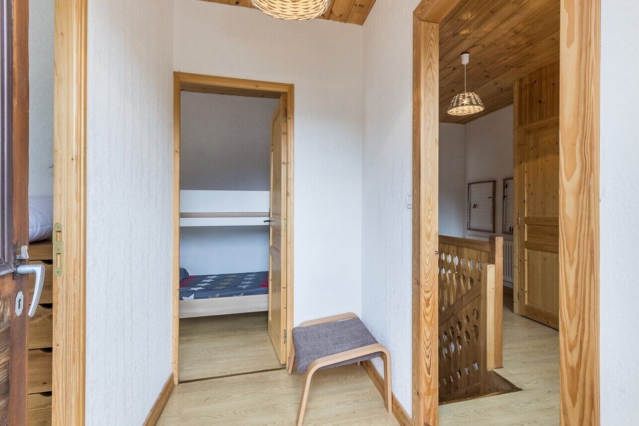 Een smalle gang met houten wanden en vloeren, die leidt naar een kamer met een stapelbed. Een klein krukje staat in de gang, en een rieten lamp hangt aan het plafond.
