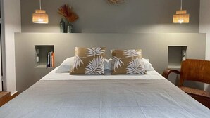 A bedroom with a large bed, two wall-mounted lamps, a decorative wall piece, and a wooden chair. - GLIGLI FOR LOVERS (Sainte-Anne)