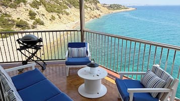 Un balcon doté de meubles bleus et blancs, d’un barbecue et offrant une vue sur la mer et les collines.