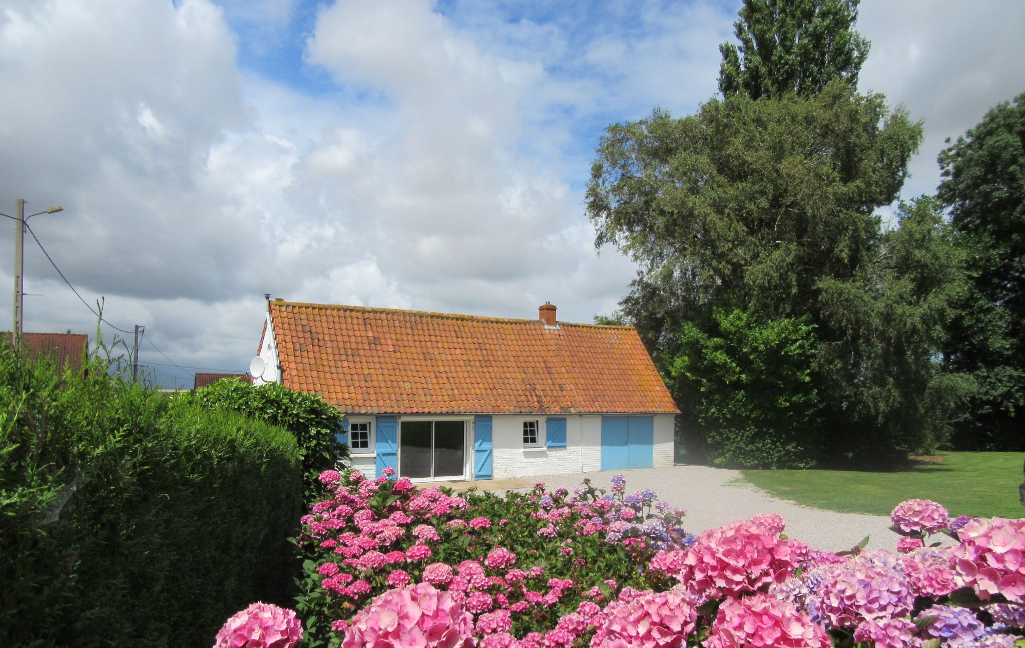 A white house with a red-tiled roof, surrounded by lush greenery and pink flowers.