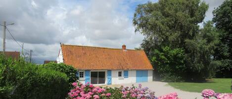 A white house with a red-tiled roof, surrounded by lush greenery and pink flowers.