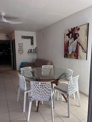 Un comedor con una mesa de vidrio rodeada de sillas blancas de patas de madera, un sofá marrón y un cuadro colgado en la pared.