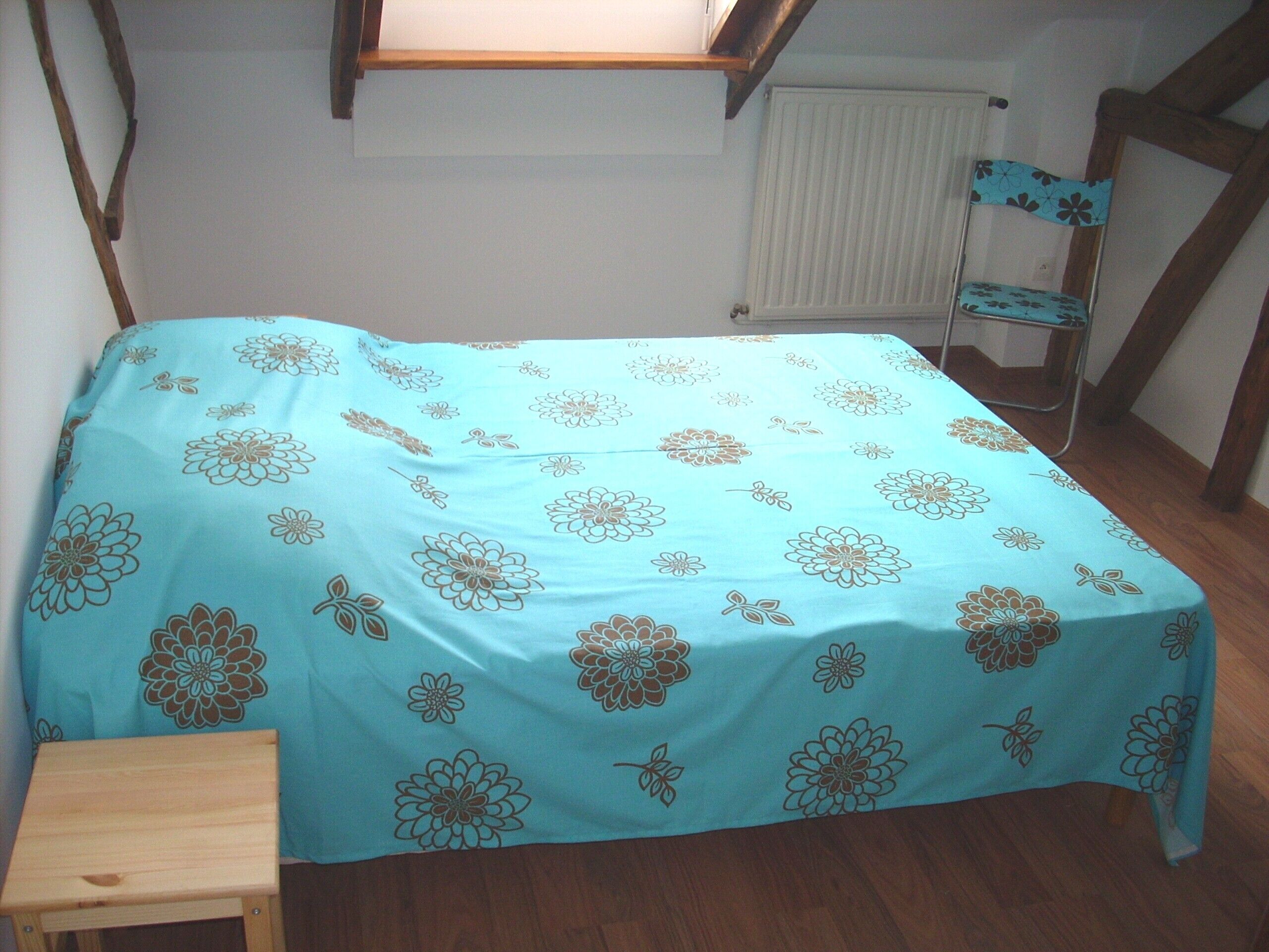A bedroom with a bed covered in a floral-patterned bedspread, a small wooden stool, and a chair with a floral cushion.