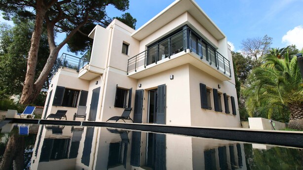 Une maison moderne à deux étages, dotée d’un balcon, de grandes fenêtres et dont le reflet se mire dans l’eau.