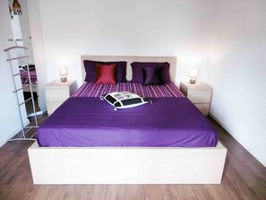 A neatly made bed with a purple bedspread and pillows, two bedside tables with lamps, and a wooden floor.