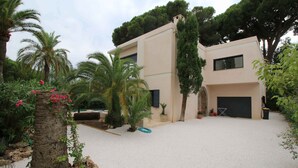 Une maison moderne à deux étages, dotée d’une allée, entourée de palmiers et de plantes en pot.