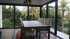 Un espace repas avec une grande table et des chaises, de grandes fenêtres donnant sur des palmiers et un luminaire suspendu.