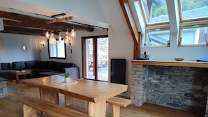 Un vaste espace de vie comprenant une table à manger en bois, des bancs, une cheminée en pierre et une grande fenêtre donnant sur un mur de pierres.