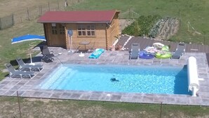 A rectangular swimming pool with a wooden deck, lounge chairs, and a small wooden cabin with a red roof.