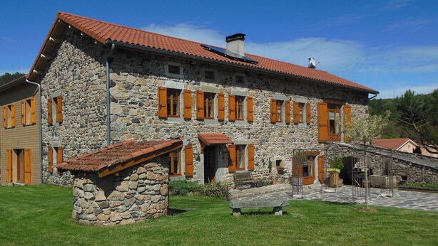 A stone building with a red-tiled roof, multiple windows with wooden shutters, and a stone bench in the foreground.