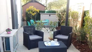 Eine Terrasse mit zwei Korbstühlen, einem kleinen Tisch, einem Blumentopf und einem Garten im Hintergrund.