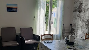 Ein Essbereich mit Tisch für zwei Personen, zwei gepolsterten Stühlen, einer wandmontierten Klimaanlage, einer Tür mit Blick auf Grünflächen und einer Wand mit einem blattgemusterten Vorhang.