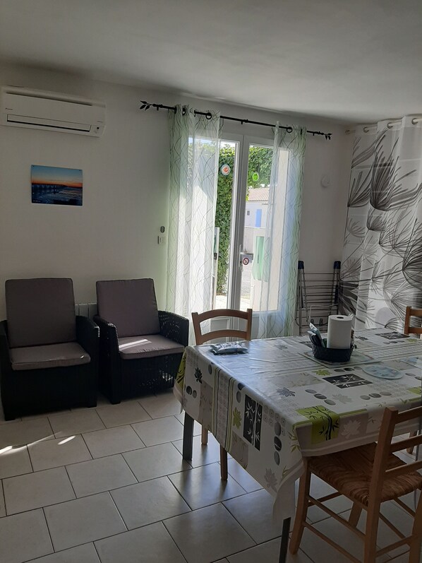 A dining area with a table set for two, two cushioned chairs, a wall-mounted air conditioner, a door with a view of greenery outside, and a wall with a leaf-patterned curtain.