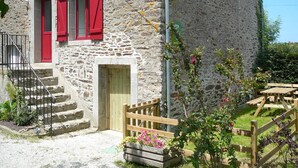 Ein Steinhaus mit roten Fensterläden, einem hölzernen Tor und einem kleinen Garten mit Blumen.