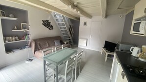 Ein kompakter Wohnraum mit Küchenzeile, einem Essbereich mit Glastisch, einer Couch, einem Bücherregal und einer Treppe zu einer oberen Ebene.