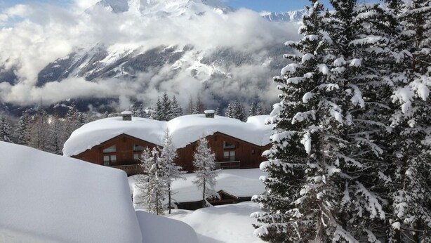 A mountain lodge with snow-covered roofs and pine trees, surrounded by snow and clouds. - South-facing apt at the foot of the ski run. View of Tarentaise valley (Montvalezan)